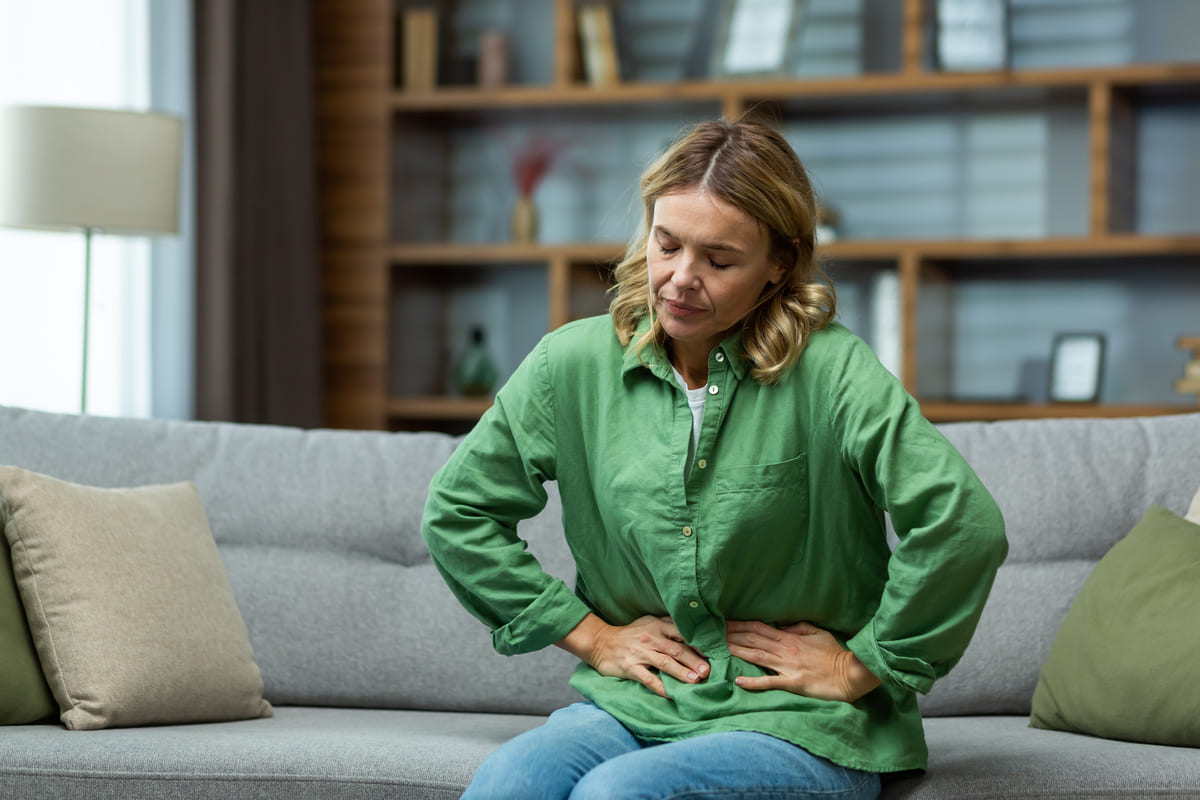 Na imagem, vemos uma mulher loira vestindo uma camisa verde, sentada em um sofá, com as mãos na barriga, indicando um possível desconforto. Ela parece estar sentindo dores abdominais, o que pode estar relacionado a condições como a síndrome do intestino irritável, uma condição que frequentemente causa dor abdominal e desconforto digestivo. A expressão dela reflete esse desconforto enquanto está em um ambiente doméstico.