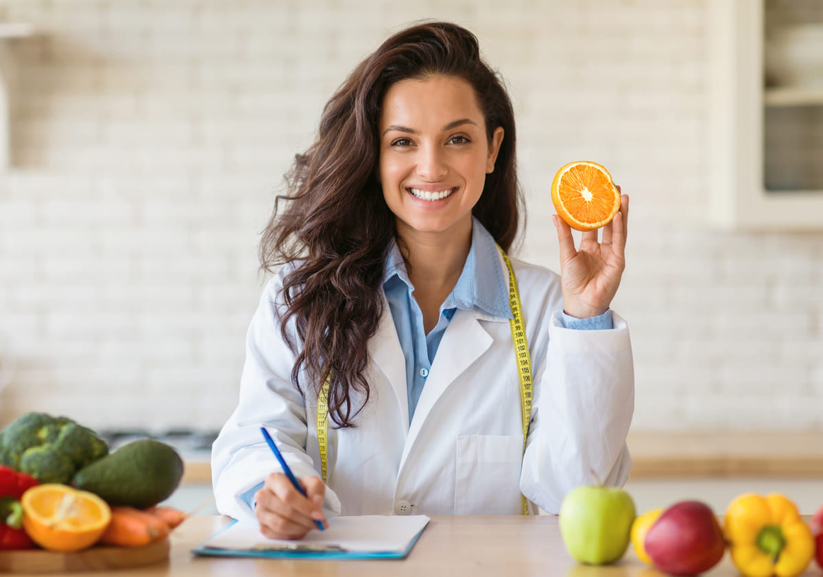 Mulher profissional da saúde alimentar segurando uma laranja e fazendo anotações, representando o nutrólogo ou nutricionista.