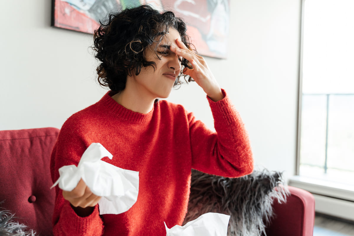 Mulher com mão no rosto e expressão de agonia, sua outra mão segura um lenço de papel, em alusão aos sintomas de uma virose.
