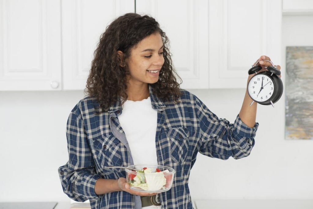 Mulher sorridente segurando uma salada e um relógio na cozinha, ilustrando o que é jejum intermitente e os horários controlados para alimentação saudável.
